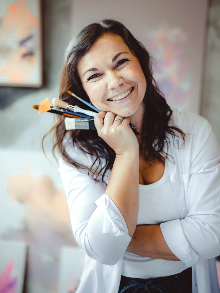 Anca Juliana Schulte – Künstlerin Künstlerin Anca Juliana Schulte hält Pinsel in der Hand, umgeben von Kunstwerken.