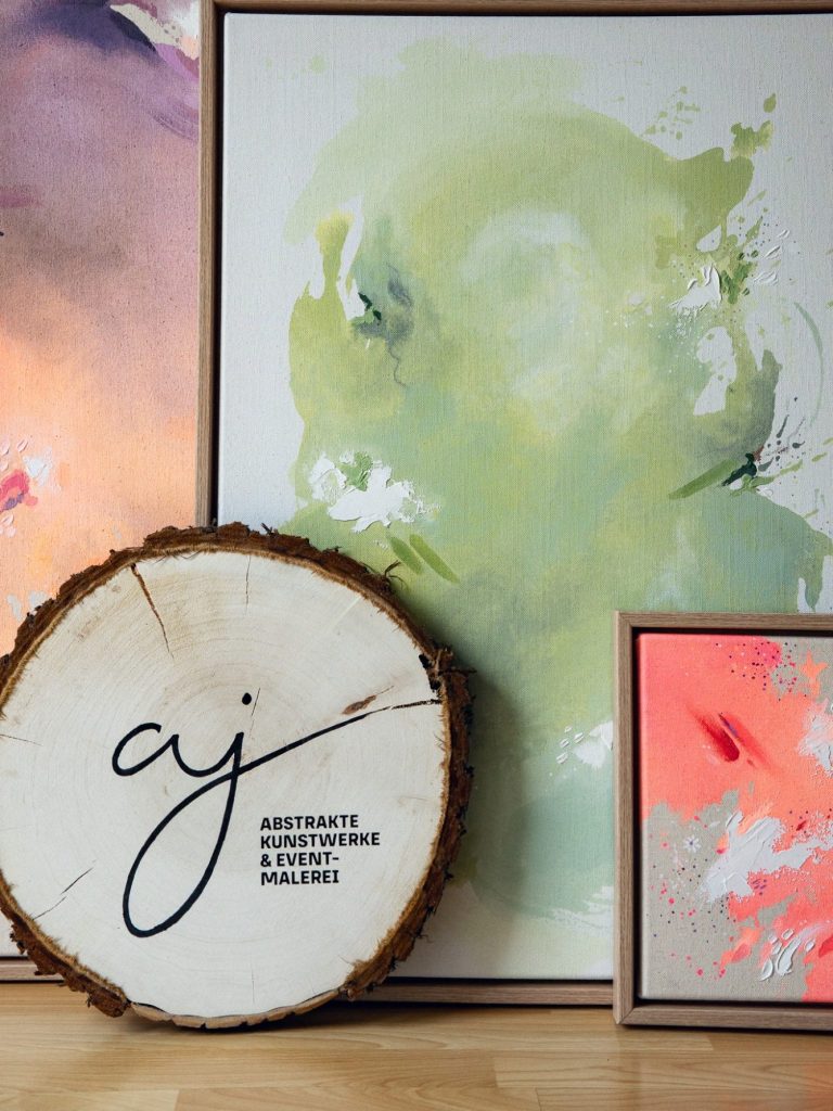 Abstrakte Kunstwerke & Event-Malerei Zwei Bilder in sanften Farben und ein Holzschild mit Logo.